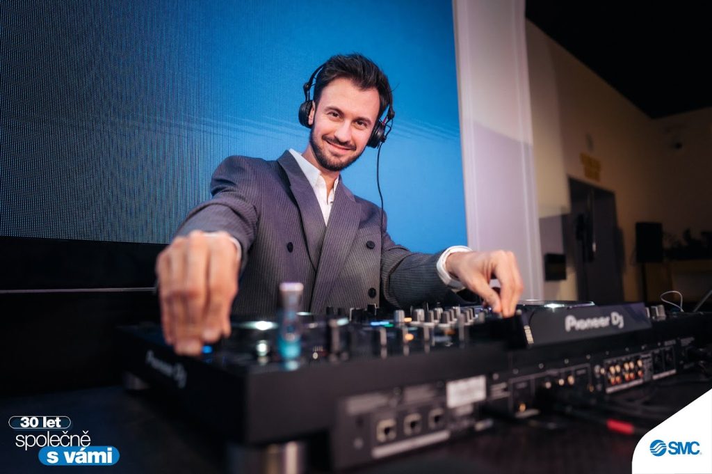 Smiling DJ Bury in a jacket with headphones behind the decks at a corporate event with an LED wall