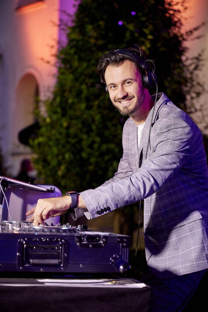 Usměvavý DJ Bury v kostkovaném saku za DJ pultem, DJ pro firemní akce a eventy