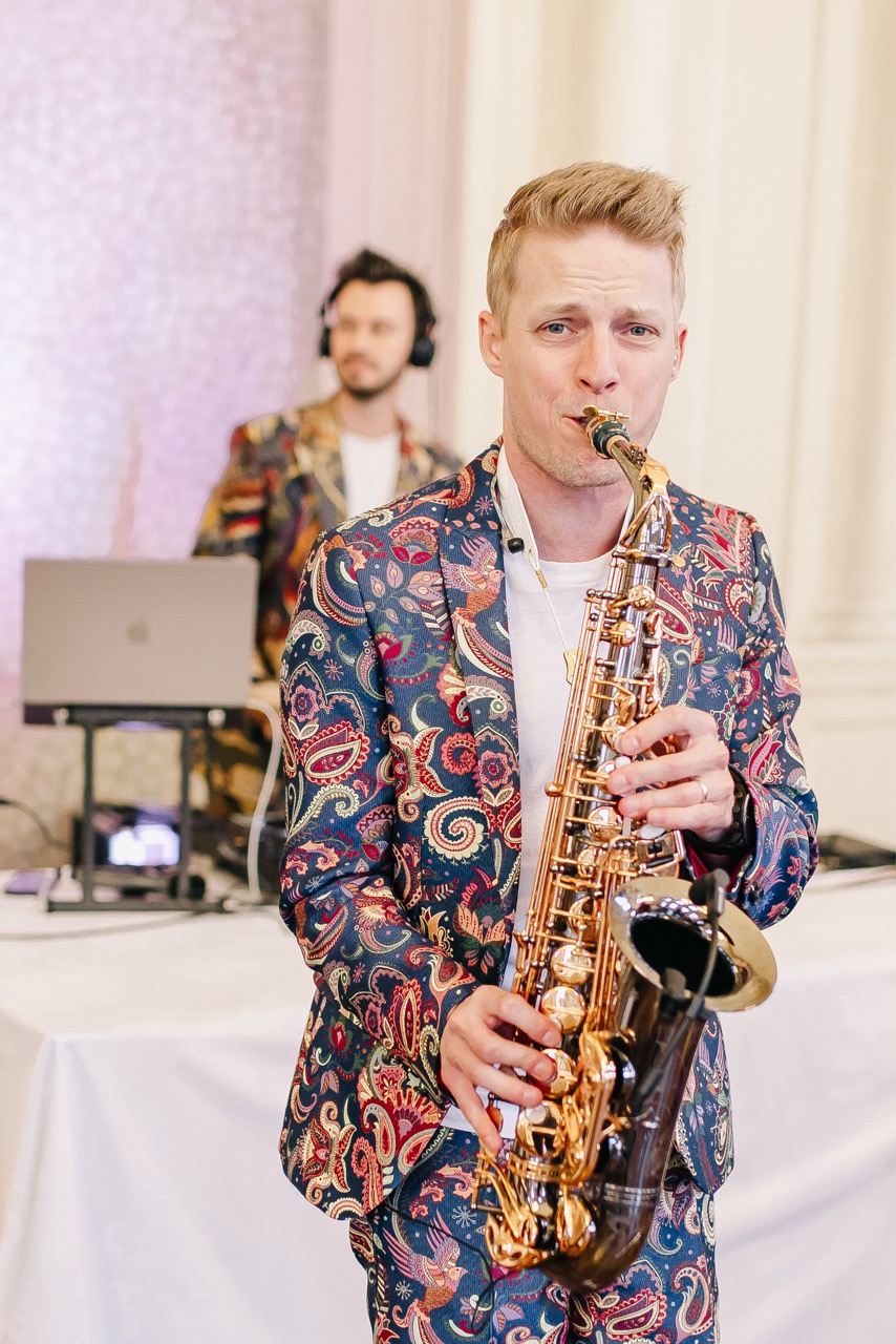 Saxofonista v květovaném saku hraje na svatební párty, za ním DJ u pultu