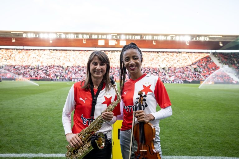 Saxofonistka Nikisaxi a houslistka Iska.muse v dresech SK Slavia Praha se saxofonem a houslemi na trávníku Eden Areny