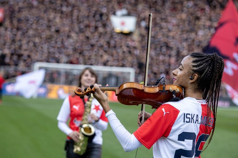 Houslistka Iska.muse v dresu Slavie hraje na housle na trávníku Eden Areny, za ní saxofonistka Nikisaxi a vlajky fanoušků